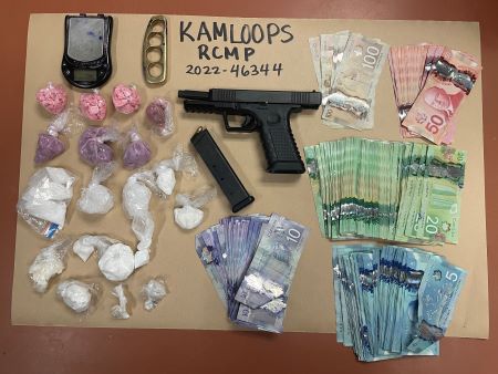 A scale, brass knuckles, black pistol, and baggies of pink, purple, and white suspected illegal drugs, in addition to stacks of Canadian currency, are placed on a sheet of brown paper with a Kamloops RCMP file number written on the top centre.
