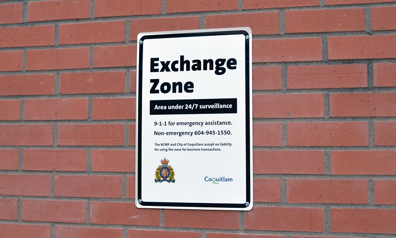 Picture of a Safe Exchange Zone sign post on brick wall states: Exchange zone Areas under 24/7 surveillance 9-1-1 for emergency assistance Non-emergency 604-945-1550 The RCMP and City of Coquitlam accept no liability for using the zone for business transactions.