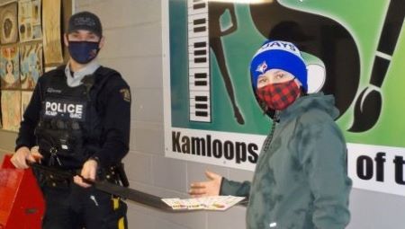 A Kamloops RCMP officer dressed in a police uniform, mask, and police ball cap uses a black goalie stick to pass white, green, and pink slurpee vouchers to a student in a green coat, red and black plaid mask, and blue and white Jays toque during a previous Random Acts of Kindness Day. 