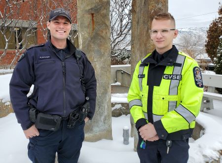 From left Kamloops RCMP Cpl. David Tunbridge, dressed in his police uniform, stands next to CVSE officer Josh Burneau, dressed in a yellow high visibility coat. Snow covers the benches and landscape behind them.