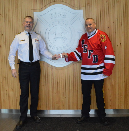 Photo of Fire Chief Warner and RCMP Superintendent Wright at Fire Hall 1. Superintendent Wright is wearing the PG Fire Fighters hockey jersey and is giving $50 to Fire Chief Warner.  