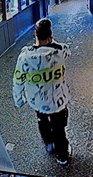 Le suspect est de dos. Il porte sa veste blanche arborant des lettres noires et une large bande jaune au milieu.