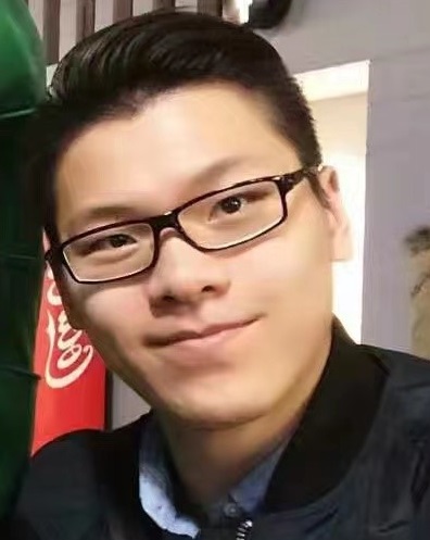 Young Asian man with black hair, brown eyes, wearing black rimmed glasses, a black zip up jacket and a blue button up dress shirt.